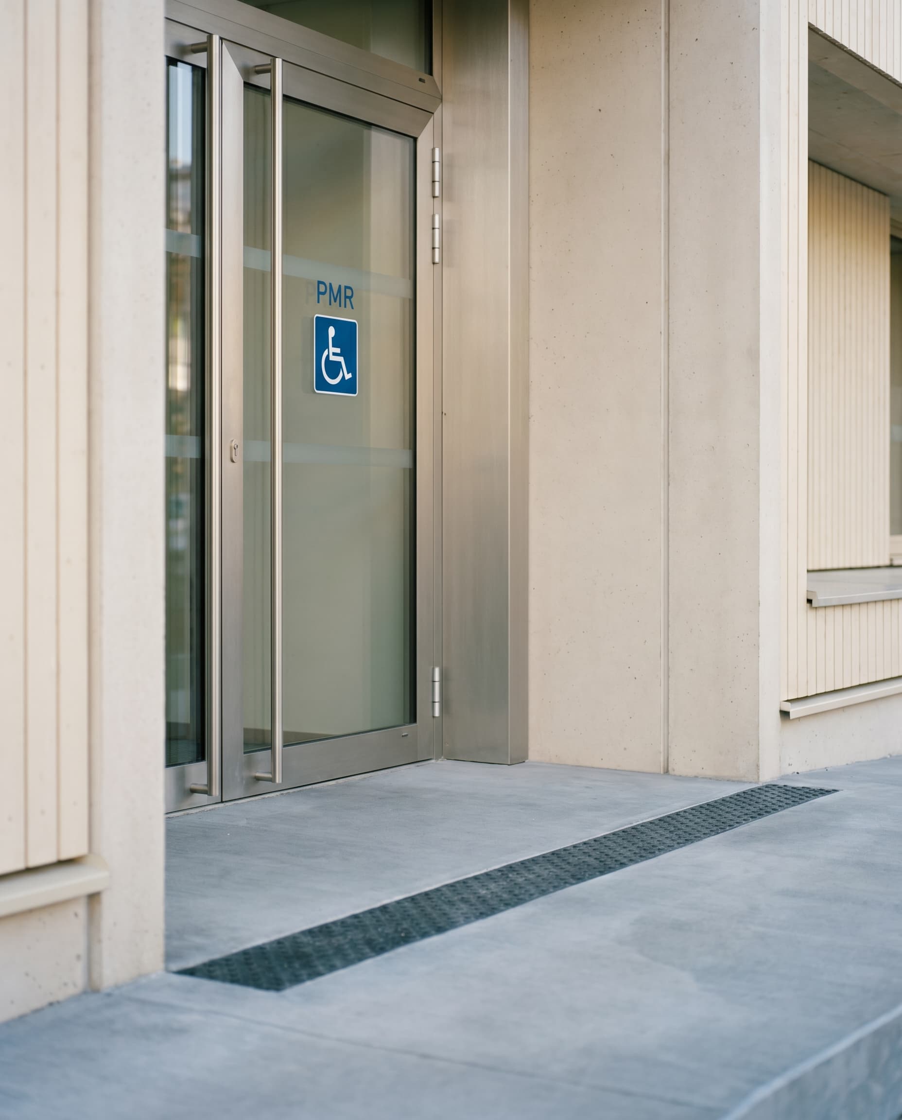 Détail d'une entrée PMR : porte vitrée avec pictogramme d'accessibilité et rampe avec bande antidérapante